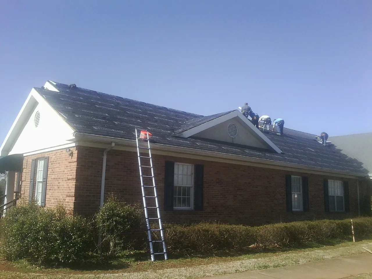 Crew tearing shingles off a brick ranch roof with a ladder set — Metal Roof Repair work by our crew in Alplaus