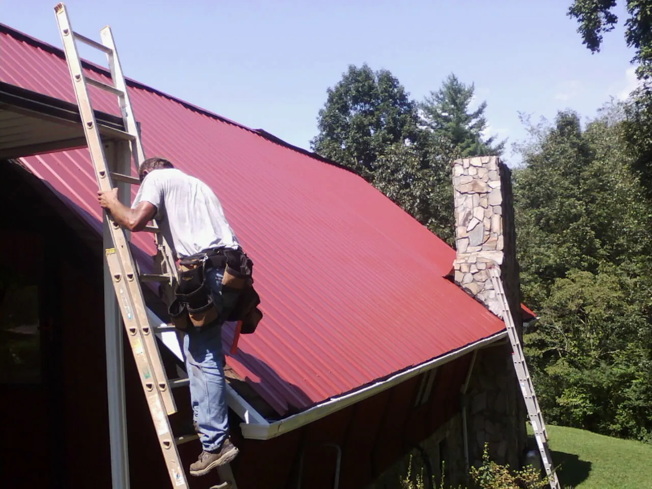 Roofer on a ladder beside a red metal roof and stone chimney — Soffit & Fascia Repair by Roofing Brothers in Alplaus