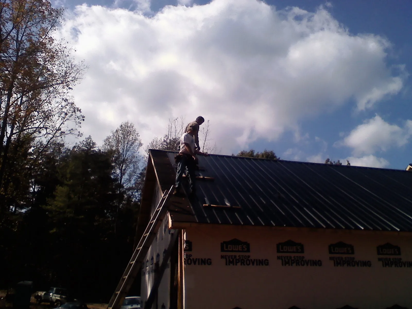 Roofer fastening dark metal panels high against a cloudy sky, from a Roof Inspection job in Alplaus